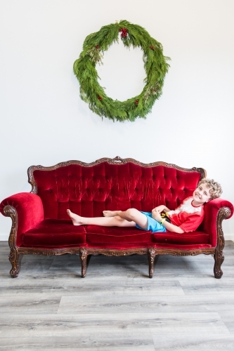 Boy sitting on red lounge smiling at Christmas with wreath - Australian Stock Image