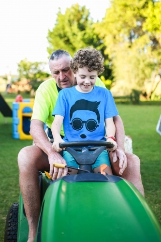 Boy sitting on ride on lawn mower with grandfather - Australian Stock Image