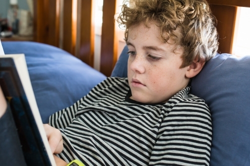 Boy sitting propped up against pillow on bed reading book natural light - Australian Stock Image