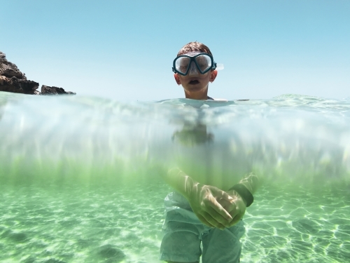 Boy standing in ocean with view underwater of body and above water of head wearing snorkel mask - Australian Stock Image
