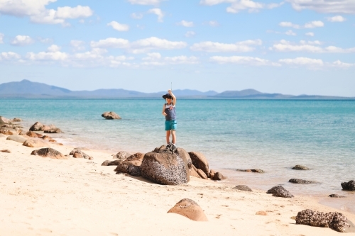 Boy standing on rocks with fishing rod on tropical beach with islands in the distance - Australian Stock Image
