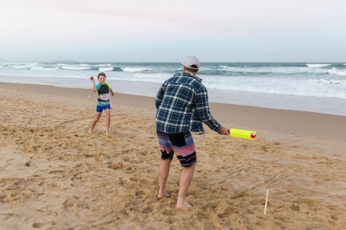 Boy throwing ball to his father holding bat playing cricket on the beach - Australian Stock Image