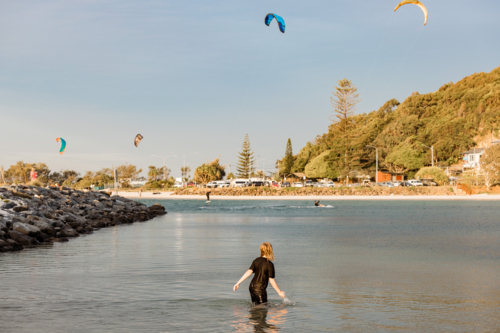 Boy wading in shallow water of Currumbin Creek watching kite surfers in the distance - Australian Stock Image