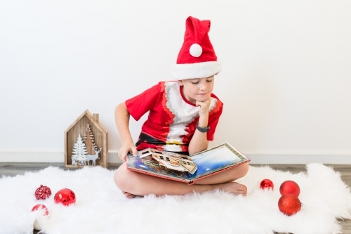 Boy wearing santa hat sitting on rug with Christmas decorations reading book - Australian Stock Image