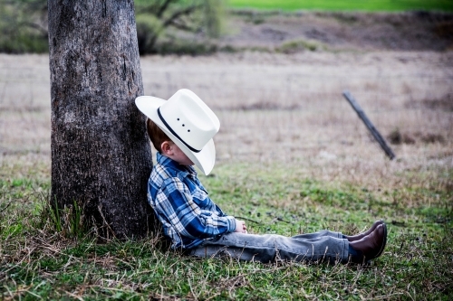 Boy with hat resting against tree on farm - Australian Stock Image