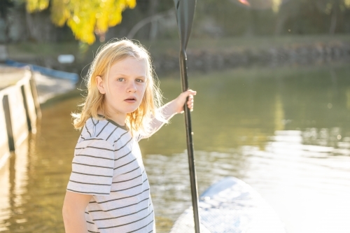 Boy with long hair standing next to lake holding SUP paddle - Australian Stock Image