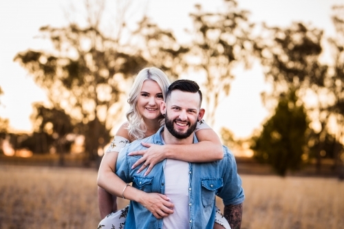 Boyfriend carrying girlfriend piggy back in paddock laughing - Australian Stock Image