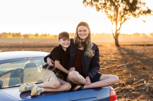 Brother and sister sitting on boot of car at sunset in dirt paddock on farm
