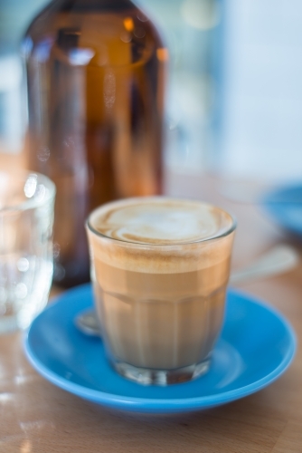 Cafe latte coffee on blue saucer with water bottle and glass - Australian Stock Image