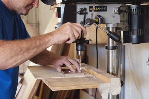 Carpenter using vertical drilling machine in woodwork workshop - Australian Stock Image