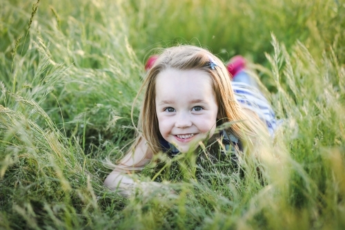 Caucasian girl with long hair lying in long grass at the park with a big smile - Australian Stock Image