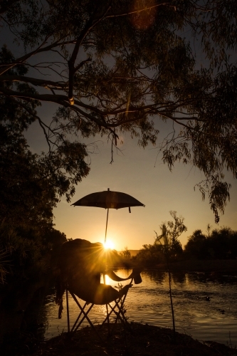 Chair with umbrella during sunrise with orange sky in front of creek with trees - Australian Stock Image