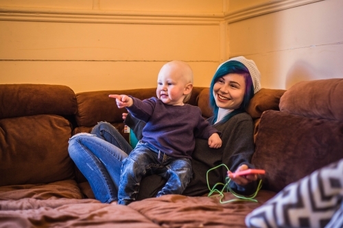 Child sitting on mum's lap on lounge pointing and smiling while mum holds phone - Australian Stock Image