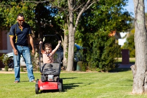 child trying to push lawnmower watched by father - Australian Stock Image