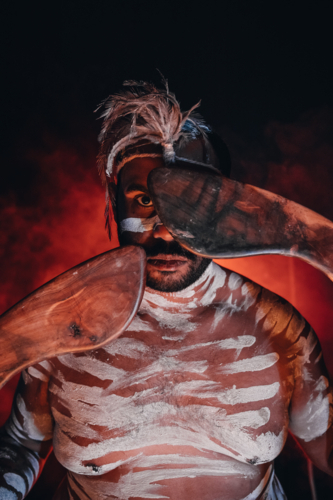 Close-up of aboriginal man holding a pair of boomerangs to his face. - Australian Stock Image