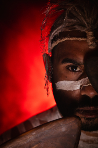 Close-up of aboriginal man holding a pair of boomerangs to his face. - Australian Stock Image