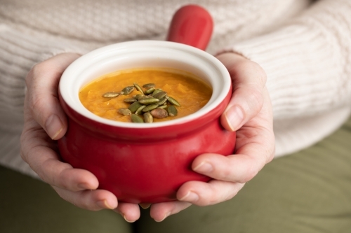 close up of hands holding red bowl of pumpkin soup - Australian Stock Image