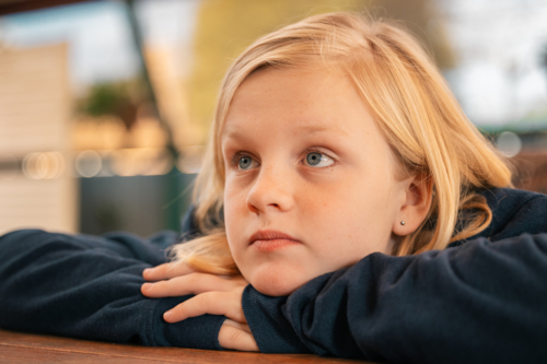 Close up portrait of pre-teen boy with long blond hair and blue eyes resting chin on hands looking - Australian Stock Image