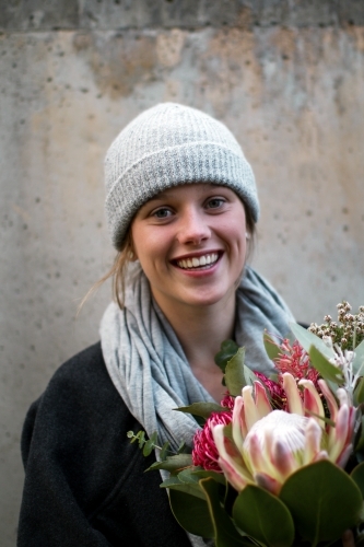Close up portrait of young girl wearing a beanie and scarf smiling whilst holding a floral bouquet - Australian Stock Image