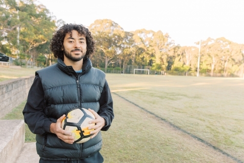 Close up shot of a smiling young man with curly hair standing on the field with a soccer ball