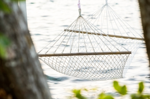 close up shot of a white string hammock near an ocean water with blurred green leaves - Australian Stock Image