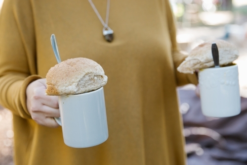 Close up shot of a woman holding two white mugs, a utensil and bread on top of each mugs - Australian Stock Image