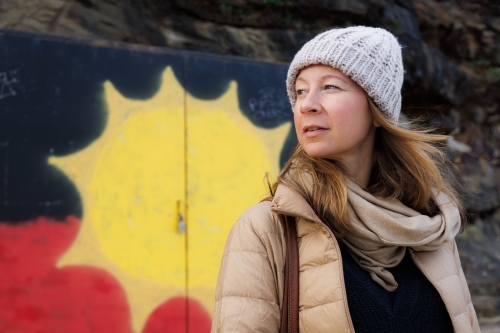 Woman wearing a beanie and coat looking away with a sun aboriginal flag in background - Australian Stock Image
