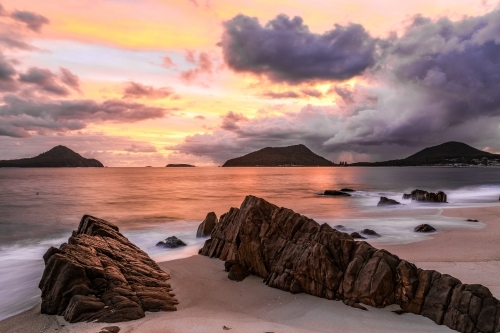 Cloudy sunrise over mountains and rocky beach - Australian Stock Image