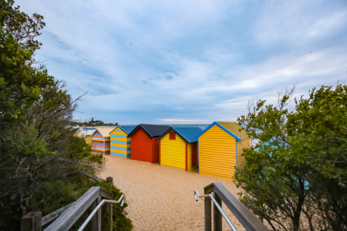 Colourful beach huts on sandy shore with greenery and cloudy sky - Australian Stock Image