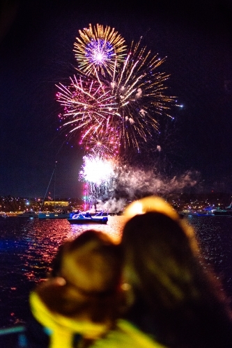 Colourful fireworks display over water with out of focus people in foreground - Australian Stock Image