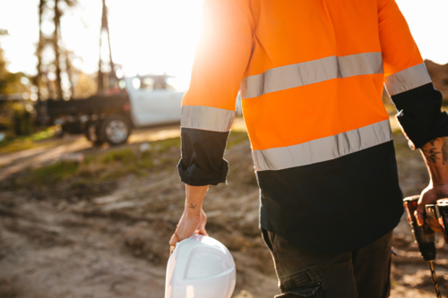 Construction worker holding a helmet and a drill while walking on the construction site. - Australian Stock Image