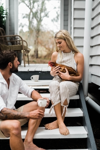 Couple having a cup of coffee on steps of verandah, with woman holding a chicken.