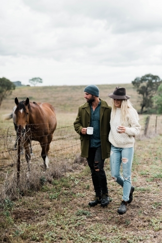 Couple walking past horses on a farm, holding cups of coffee - Australian Stock Image