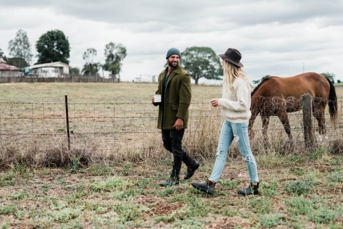 Couple walking past horses on a farm, holding cups of coffee