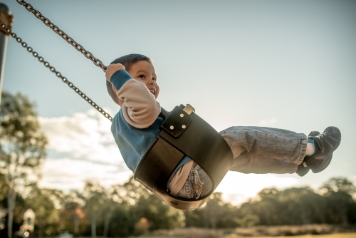 Cute mixed race three year old boy playing on a swing in a suburban playground - Australian Stock Image