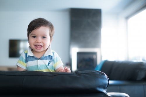 Cute mixed race toddler plays on a black leather lounge at home - Australian Stock Image