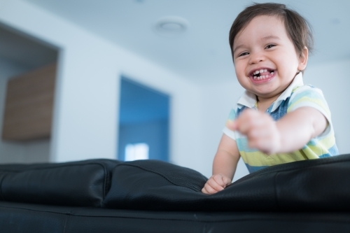 Cute mixed race toddler plays on a black leather lounge at home - Australian Stock Image