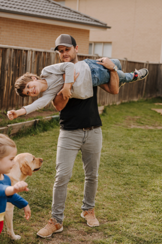 Dad holding his son upside down pretending to fly playing together in backyard outside home - Australian Stock Image