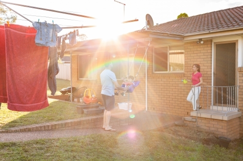 Dad pushing toddler boy on swing in backyard with sun flare - Australian Stock Image