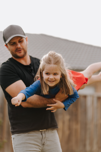 Dad simulating a flight for daughter in superhero costume. - Australian Stock Image