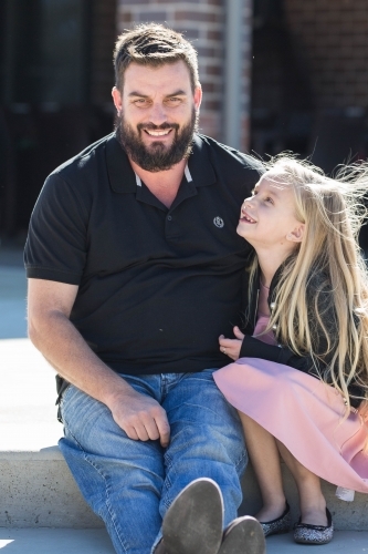 Daughter looking up at father sitting together outside - Australian Stock Image