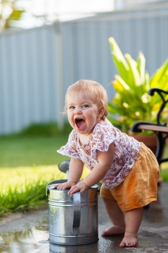 Delighted baby with huge smile discovering water in garden watering can - Australian Stock Image