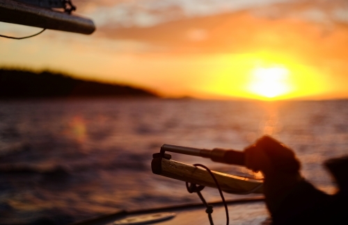 Detail of rudder and skipper's hand on small sailing boat in vast seascape at sunset - Australian Stock Image