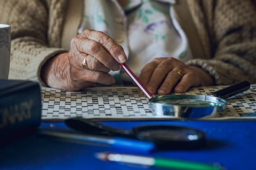 Elderly lady's hands with crossword puzzle, magnifying glass, pencil, wedding ring, and dictionary