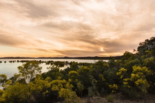 Expansive sky at sunset over large dam of water with wattle trees - Australian Stock Image
