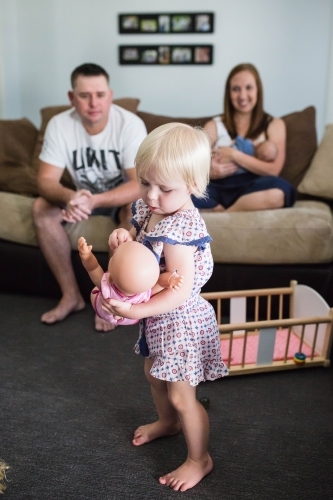 Family at home sitting on lounge with mother feeding newborn baby watching daughter play with doll - Australian Stock Image