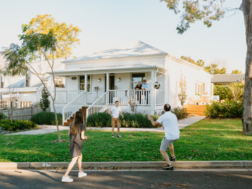 Family playing soccer in front yard of white weatherboard home in suburbs - Australian Stock Image