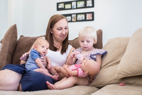 Family sitting on lounge with mum burpring newborn son while daughter plays with doll