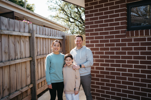 Family standing in front of the brick wall of their house. - Australian Stock Image