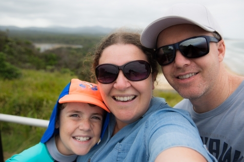 Family standing together happy smiling taking selfie with beach and trees behind - Australian Stock Image